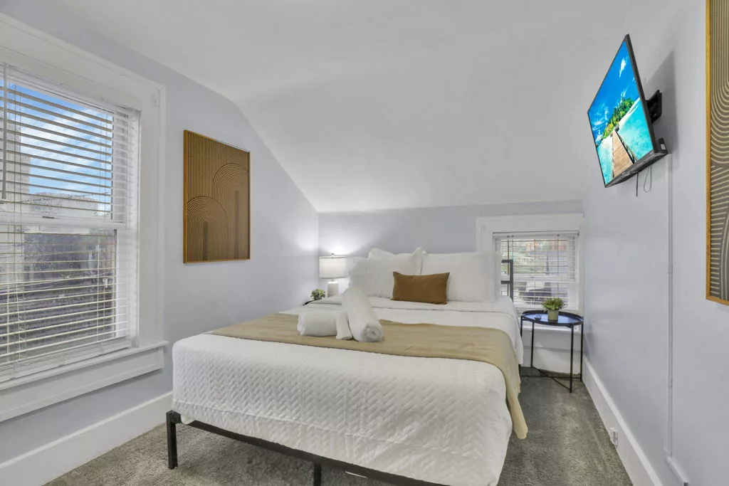 The Lantern bedroom with white bedding, a gold throw, framed artwork, windows, and a wall-mounted television.