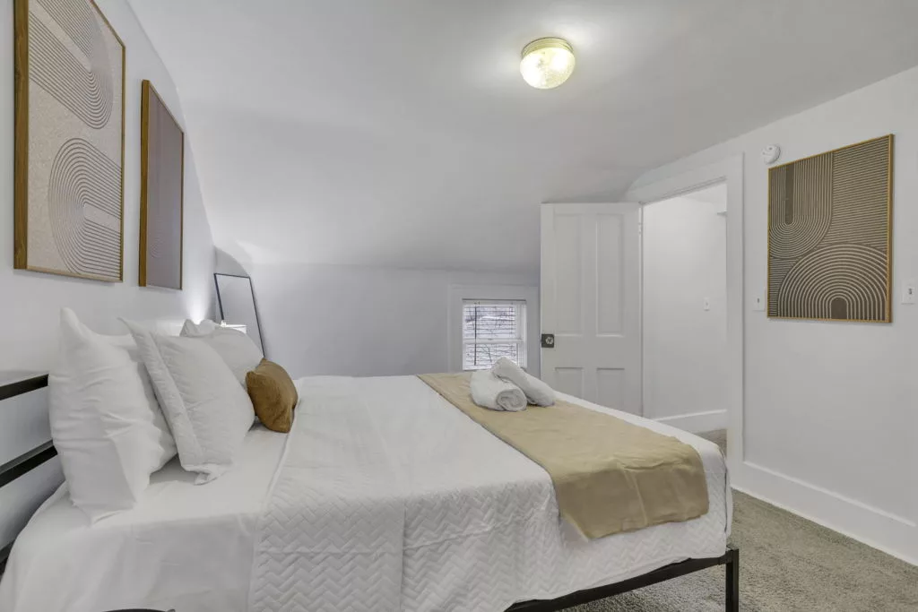 The Lantern second bedroom with white bedding, a gold throw, geometric artwork, and a sloped ceiling.