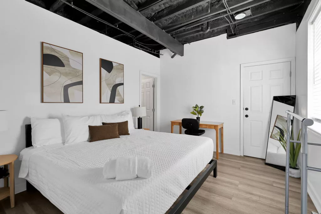King bedroom with white bedding, a wood desk and black chair, a full-length mirror, and a clothes rack beside the window.