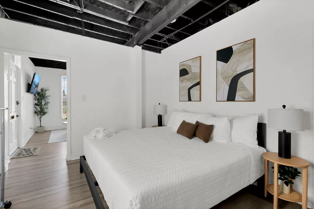King bed with white bedding, warm brown pillows, abstract artwork, and matching bedside lamps beneath exposed black ceiling beams.