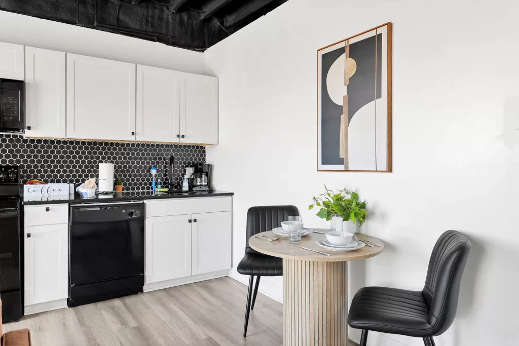 Round wood dining table set for two with black chairs, a green plant, and abstract artwork beside the kitchen.